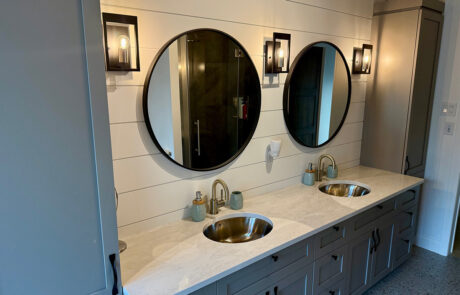 Photo d’une salle de bain neuve avec vanité double : comptoir en quartz pâle, lavabos ronds en inox, robinetterie laiton, deux miroirs ronds noirs et appliques murales
