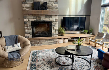 Photo d’un salon neuf avec foyer en pierre et manteau de bois, télévision murale sur long banc, tables basses rondes noires, fauteuil d’appoint et tapis à motifs