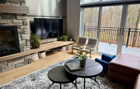 Photo d’un salon lumineux avec foyer en pierre, long banc TV en bois, plantes, grandes portes-patio donnant sur la terrasse, tables basses rondes noires et tapis à motifs