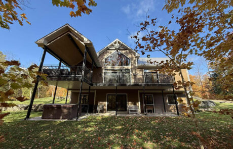 Photo de la façade arrière d’une maison unifamiliale neuve avec grande terrasse couverte sur poteaux, garde-corps noirs, spa au rez-de-jardin et feuillage d’automne